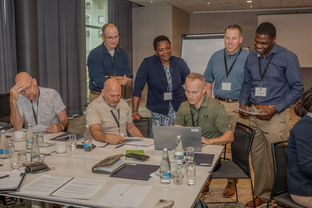 A group of approximately eight professionals are gathered around a conference table in what appears to be a hotel meeting room or corporate training facility. The group is focused on a laptop screen, suggesting a collaborative work session or workshop. Several participants are wearing lanyards with name badges, indicating a formal professional event.