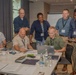 A group of approximately eight professionals are gathered around a conference table in what appears to be a hotel meeting room or corporate training facility. The group is focused on a laptop screen, suggesting a collaborative work session or workshop. Several participants are wearing lanyards with name badges, indicating a formal professional event.