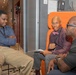 Three men are engaged in what appears to be a professional conversation or interview in a modern office or hotel lobby setting. The man in the center, wearing a purple shirt and pink bow tie with glasses, holds a notepad and pen, suggesting he may be conducting an interview or taking notes. The setting features distinctive orange chairs and contemporary interior design.