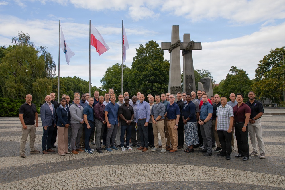 A large group of approximately 40 adults poses for a formal group photograph in front of a prominent monument featuring three large stone crosses bound together with rope. The Polish national flag is visible among the flagpoles, and the setting appears to be a public memorial square with cobblestone paving. The monument is consistent in appearance with the Three Crosses Monument (Pomnik Trzech Krzyży) in Poznań, Poland, commemorating the 1956 Poznań protests.