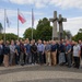 A large group of approximately 40 adults poses for a formal group photograph in front of a prominent monument featuring three large stone crosses bound together with rope. The Polish national flag is visible among the flagpoles, and the setting appears to be a public memorial square with cobblestone paving. The monument is consistent in appearance with the Three Crosses Monument (Pomnik Trzech Krzyży) in Poznań, Poland, commemorating the 1956 Poznań protests.