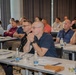 A group of approximately 20-30 adult men are seated at white tables in a conference or training room setting. The attendees appear to be engaged in listening to a presentation or lecture, with notebooks, water bottles, and glasses on the tables. The setting suggests a professional seminar, workshop, or corporate training event.