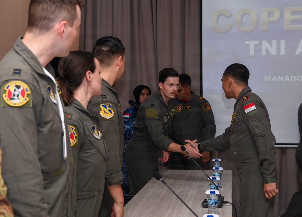 Military personnel in olive/sage green flight suits are gathered in a conference room for what appears to be a joint military exercise briefing. A person wearing an Indonesian flag patch on their right shoulder is shaking hands with a US Air Force member, suggesting a bilateral military cooperation event. The presentation screen in the background displays partial text reading 'COPE' and 'TNI A' with 'MANADO' visible, indicating this is likely a 'Cope' series joint exercise involving the Indonesian Air Force (TNI AU) in Manado, Indonesia.