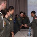 Military personnel in olive/sage green flight suits are gathered in a conference room for what appears to be a joint military exercise briefing. A person wearing an Indonesian flag patch on their right shoulder is shaking hands with a US Air Force member, suggesting a bilateral military cooperation event. The presentation screen in the background displays partial text reading 'COPE' and 'TNI A' with 'MANADO' visible, indicating this is likely a 'Cope' series joint exercise involving the Indonesian Air Force (TNI AU) in Manado, Indonesia.