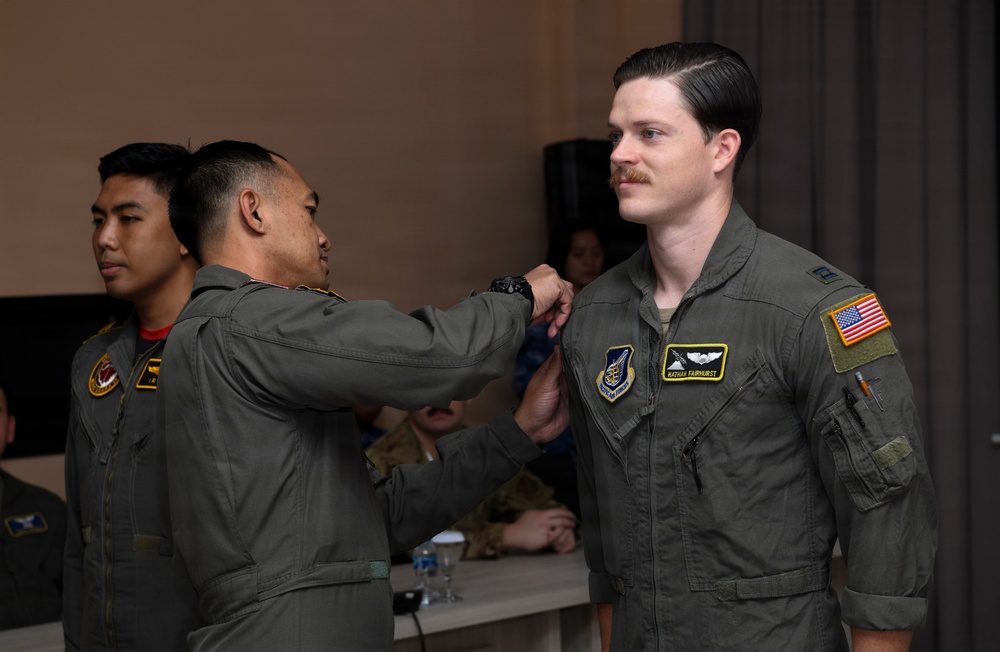 A military ceremony appears to be taking place in an indoor setting, where one service member is pinning something onto the chest of another. The recipient, whose name tag reads 'NATHAN FAIRHURST,' is wearing a U.S. Air Force flight suit with an American flag patch and pilot wings. A third service member stands in the background, also in a flight suit.
