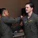 A military ceremony appears to be taking place in an indoor setting, where one service member is pinning something onto the chest of another. The recipient, whose name tag reads 'NATHAN FAIRHURST,' is wearing a U.S. Air Force flight suit with an American flag patch and pilot wings. A third service member stands in the background, also in a flight suit.
