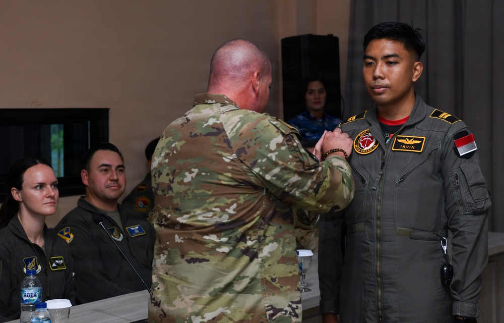 A military ceremony appears to be taking place in which a U.S. service member in OCP (Operational Camouflage Pattern) uniform is pinning a badge or award onto an Indonesian military aviator named 'Irvin,' identifiable by the Indonesian flag patch and name tape on his flight suit. Several other military personnel in flight suits are seated in the background observing the ceremony. The setting appears to be an indoor meeting or conference room.