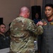 A military ceremony appears to be taking place in which a U.S. service member in OCP (Operational Camouflage Pattern) uniform is pinning a badge or award onto an Indonesian military aviator named 'Irvin,' identifiable by the Indonesian flag patch and name tape on his flight suit. Several other military personnel in flight suits are seated in the background observing the ceremony. The setting appears to be an indoor meeting or conference room.