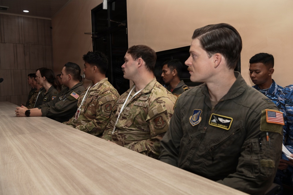 A group of military personnel from multiple nations are seated at a long table, appearing to attend a briefing or meeting. The group includes U.S. Air Force members in both OCP (Operational Camouflage Pattern) uniforms and olive green flight suits, alongside personnel in what appear to be Southeast Asian military uniforms. The setting appears to be an indoor conference or meeting room.