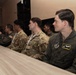 A group of military personnel from multiple nations are seated at a long table, appearing to attend a briefing or meeting. The group includes U.S. Air Force members in both OCP (Operational Camouflage Pattern) uniforms and olive green flight suits, alongside personnel in what appear to be Southeast Asian military uniforms. The setting appears to be an indoor conference or meeting room.