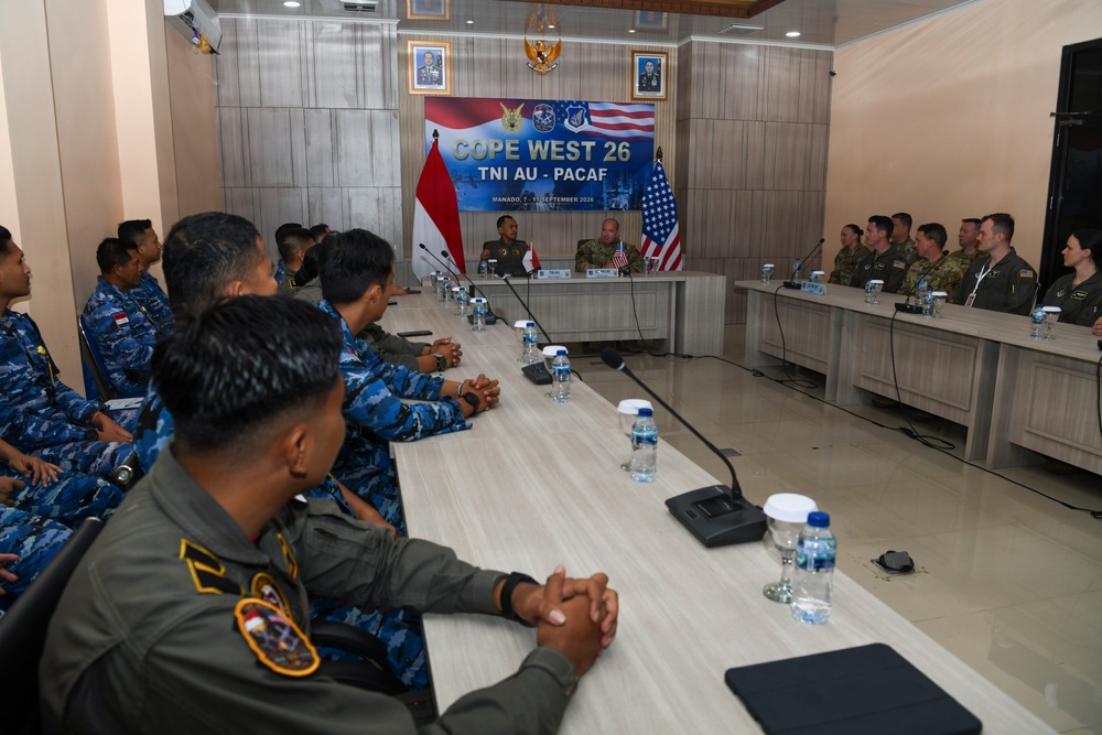 A joint military briefing or meeting is taking place for 'Cope West 26,' a bilateral exercise between TNI AU (Indonesian Air Force) and PACAF (Pacific Air Forces, U.S.), held in Manado, Indonesia in September 2026. Personnel from both nations are seated at a long conference table facing two senior officers at the head of the room. Indonesian military personnel in blue camouflage uniforms are seated on the left, while U.S. military personnel in tan/green uniforms are seated on the right.