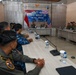 A joint military briefing or meeting is taking place for 'Cope West 26,' a bilateral exercise between TNI AU (Indonesian Air Force) and PACAF (Pacific Air Forces, U.S.), held in Manado, Indonesia in September 2026. Personnel from both nations are seated at a long conference table facing two senior officers at the head of the room. Indonesian military personnel in blue camouflage uniforms are seated on the left, while U.S. military personnel in tan/green uniforms are seated on the right.