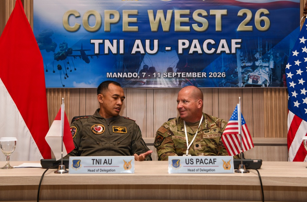 Two military officers — one from the Indonesian Air Force (TNI AU) and one from the U.S. Air Force (PACAF) — are seated at a delegation table during the Cope West 26 bilateral exercise in Manado, Indonesia, scheduled for September 7–11, 2026. Both are identified as Heads of Delegation via nameplates on the table. They appear to be engaged in a conversation, likely during an opening or planning conference.