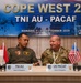 Two military officers — one from the Indonesian Air Force (TNI AU) and one from the U.S. Air Force (PACAF) — are seated at a delegation table during the Cope West 26 bilateral exercise in Manado, Indonesia, scheduled for September 7–11, 2026. Both are identified as Heads of Delegation via nameplates on the table. They appear to be engaged in a conversation, likely during an opening or planning conference.