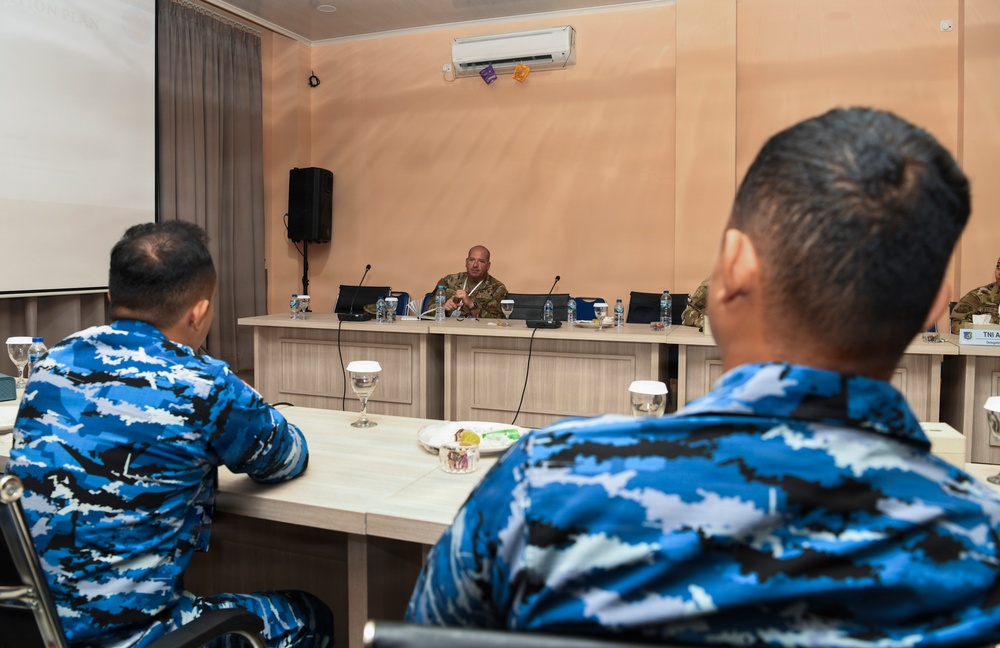 A military briefing or meeting is taking place in a conference room, with a speaker in military uniform seated at a front podium/table addressing attendees. Two individuals in the foreground are wearing distinctive blue digital camouflage uniforms, consistent with Indonesian Air Force (TNI AU) uniforms, as suggested by a partially visible nameplate. The setting appears to be a formal military exchange or joint training event.