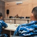 A military briefing or meeting is taking place in a conference room, with a speaker in military uniform seated at a front podium/table addressing attendees. Two individuals in the foreground are wearing distinctive blue digital camouflage uniforms, consistent with Indonesian Air Force (TNI AU) uniforms, as suggested by a partially visible nameplate. The setting appears to be a formal military exchange or joint training event.