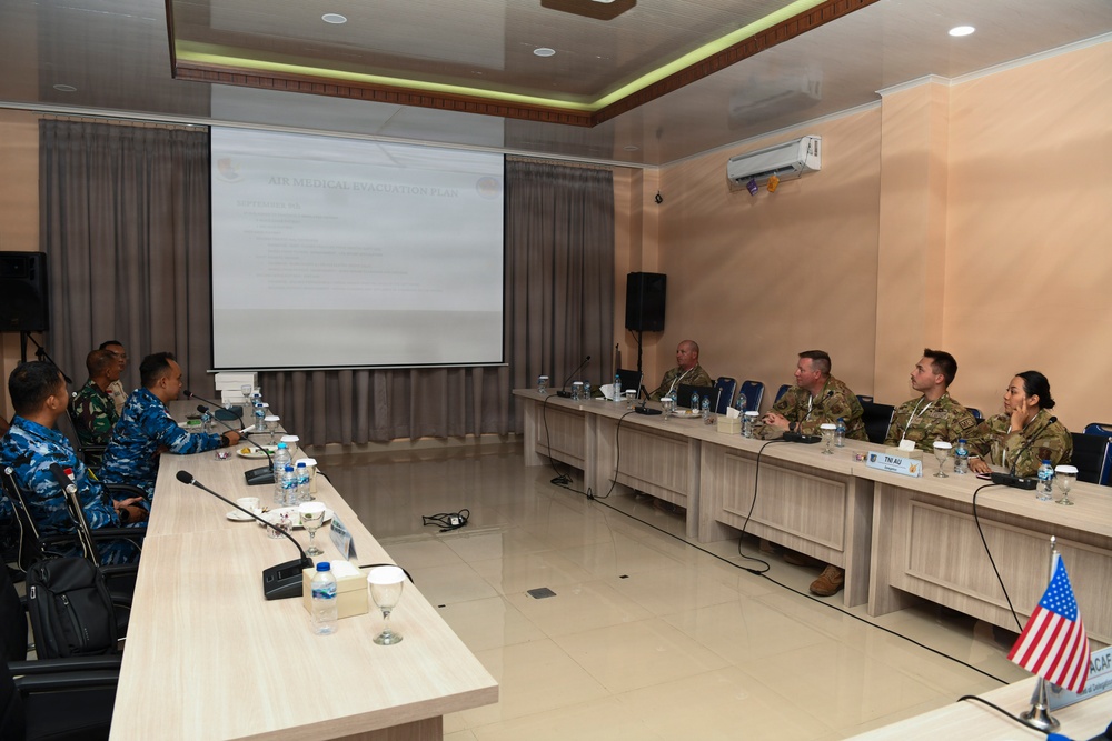 A multinational military briefing or planning meeting is taking place in a conference room, with personnel from what appears to be Indonesian military (TNI AU) and U.S. military (PACAF) seated at arranged tables. A presentation titled 'Air Medical Evacuation Plan' is displayed on a projection screen at the front of the room. The setting suggests a bilateral or multilateral military cooperation exercise or planning session.