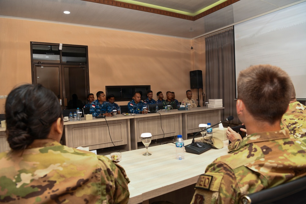 A formal military meeting or briefing is taking place in a conference room between two groups of military personnel. One group, seated at the far side of the table, wears blue naval uniforms with Indonesian flag patches, while the other group in the foreground wears green/tan camouflage uniforms consistent with Western military forces. The setting suggests a bilateral or multinational military coordination meeting.