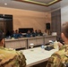 A formal military meeting or briefing is taking place in a conference room between two groups of military personnel. One group, seated at the far side of the table, wears blue naval uniforms with Indonesian flag patches, while the other group in the foreground wears green/tan camouflage uniforms consistent with Western military forces. The setting suggests a bilateral or multinational military coordination meeting.