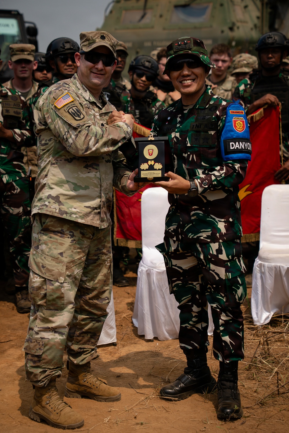 Two military officers, one wearing a U.S. Army uniform with an American flag patch and Airborne tab, and another wearing Indonesian Army (TNI-AD) camouflage with a 'DANYON' armband, are jointly holding a commemorative plaque or award. The exchange appears to be part of a formal military ceremony, likely a joint exercise or bilateral military cooperation event. A group of soldiers from both nations stands in the background near what appears to be a military helicopter.