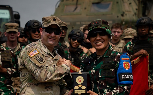 Two military officers, one wearing a U.S. Army uniform with an American flag patch and Airborne tab, and another wearing Indonesian Army (TNI-AD) camouflage with a 'DANYON' armband, are jointly holding a commemorative plaque or award. The exchange appears to be part of a formal military ceremony, likely a joint exercise or bilateral military cooperation event. A group of soldiers from both nations stands in the background near what appears to be a military helicopter.