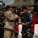 Two military officers, one wearing a U.S. Army uniform with an American flag patch and Airborne tab, and another wearing Indonesian Army (TNI-AD) camouflage with a 'DANYON' armband, are jointly holding a commemorative plaque or award. The exchange appears to be part of a formal military ceremony, likely a joint exercise or bilateral military cooperation event. A group of soldiers from both nations stands in the background near what appears to be a military helicopter.