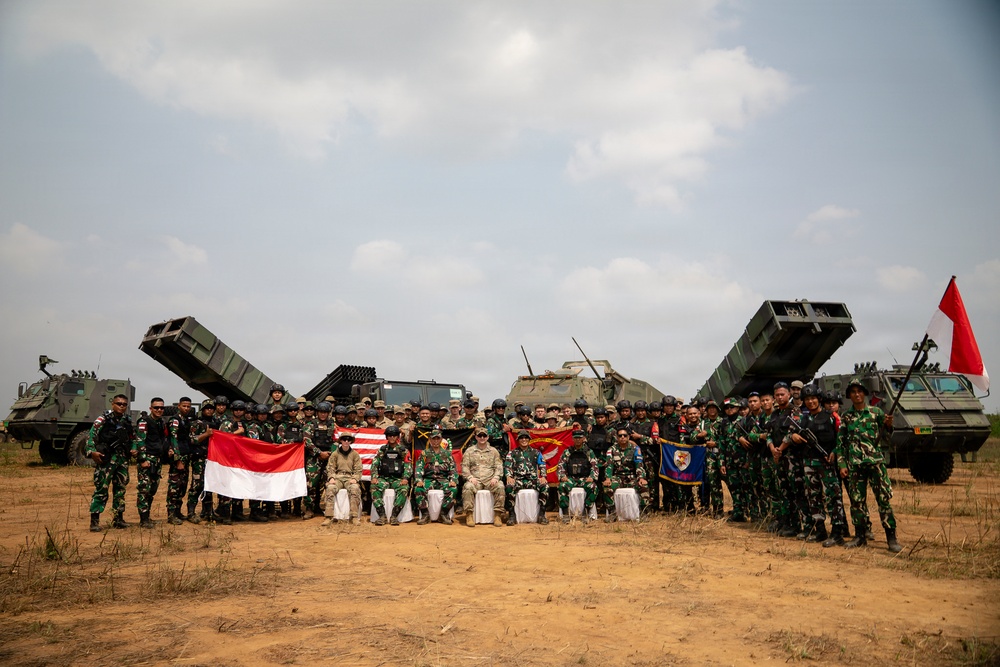 A joint military group photograph featuring personnel from Indonesia and the United States posing in front of multiple military vehicles, including what appear to be multiple rocket launcher systems (MLRS). Participants are holding Indonesian (red and white) and American flags, along with what appears to be a unit guidon. The setting is an open sandy field, suggesting a military exercise or training event.