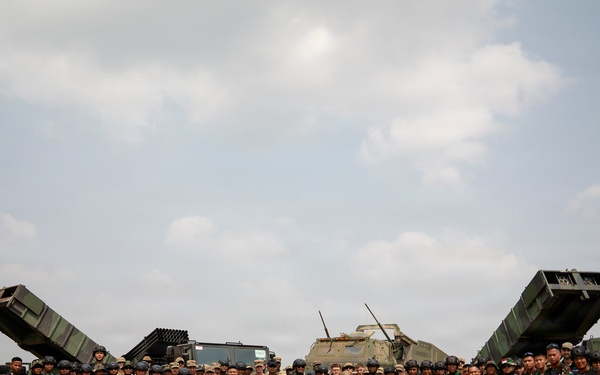 A joint military group photograph featuring personnel from Indonesia and the United States posing in front of multiple military vehicles, including what appear to be multiple rocket launcher systems (MLRS). Participants are holding Indonesian (red and white) and American flags, along with what appears to be a unit guidon. The setting is an open sandy field, suggesting a military exercise or training event.