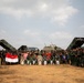A joint military group photograph featuring personnel from Indonesia and the United States posing in front of multiple military vehicles, including what appear to be multiple rocket launcher systems (MLRS). Participants are holding Indonesian (red and white) and American flags, along with what appears to be a unit guidon. The setting is an open sandy field, suggesting a military exercise or training event.