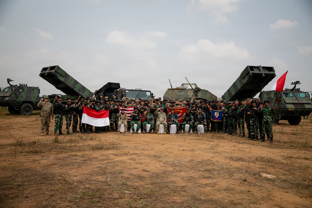 A large group of military personnel from at least two nations pose for a group photograph on an open dirt field. The group includes soldiers in camouflage uniforms alongside what appear to be American military advisors, with multiple rocket launcher systems deployed behind them. Indonesian and American flags are visibly held by participants, suggesting a joint military exercise or training event.