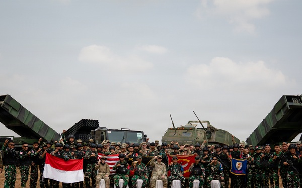 A large group of military personnel from at least two nations pose for a group photograph on an open dirt field. The group includes soldiers in camouflage uniforms alongside what appear to be American military advisors, with multiple rocket launcher systems deployed behind them. Indonesian and American flags are visibly held by participants, suggesting a joint military exercise or training event.