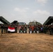 A large group of military personnel from at least two nations pose for a group photograph on an open dirt field. The group includes soldiers in camouflage uniforms alongside what appear to be American military advisors, with multiple rocket launcher systems deployed behind them. Indonesian and American flags are visibly held by participants, suggesting a joint military exercise or training event.