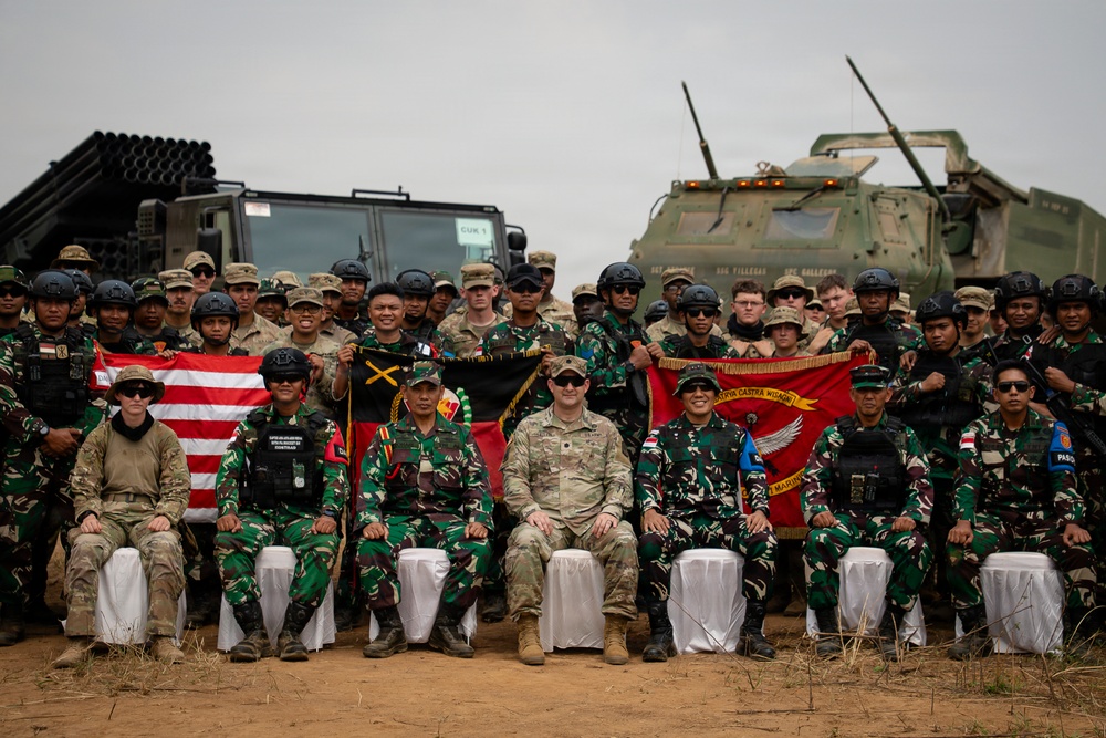 A joint military group photograph featuring soldiers from at least two nations, identifiable by distinct camouflage uniforms — Indonesian Armed Forces (TNI) in woodland pattern and U.S. Army personnel in MultiCam/OCP. Participants are posed in front of military vehicles including what appears to be a HIMARS rocket artillery system and an armored vehicle. Several unit flags and national flags are being displayed, suggesting a bilateral or multinational military exercise.