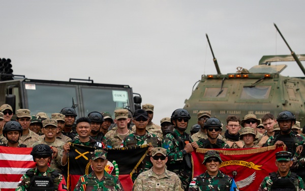 A joint military group photograph featuring soldiers from at least two nations, identifiable by distinct camouflage uniforms — Indonesian Armed Forces (TNI) in woodland pattern and U.S. Army personnel in MultiCam/OCP. Participants are posed in front of military vehicles including what appears to be a HIMARS rocket artillery system and an armored vehicle. Several unit flags and national flags are being displayed, suggesting a bilateral or multinational military exercise.