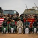 A joint military group photograph featuring soldiers from at least two nations, identifiable by distinct camouflage uniforms — Indonesian Armed Forces (TNI) in woodland pattern and U.S. Army personnel in MultiCam/OCP. Participants are posed in front of military vehicles including what appears to be a HIMARS rocket artillery system and an armored vehicle. Several unit flags and national flags are being displayed, suggesting a bilateral or multinational military exercise.