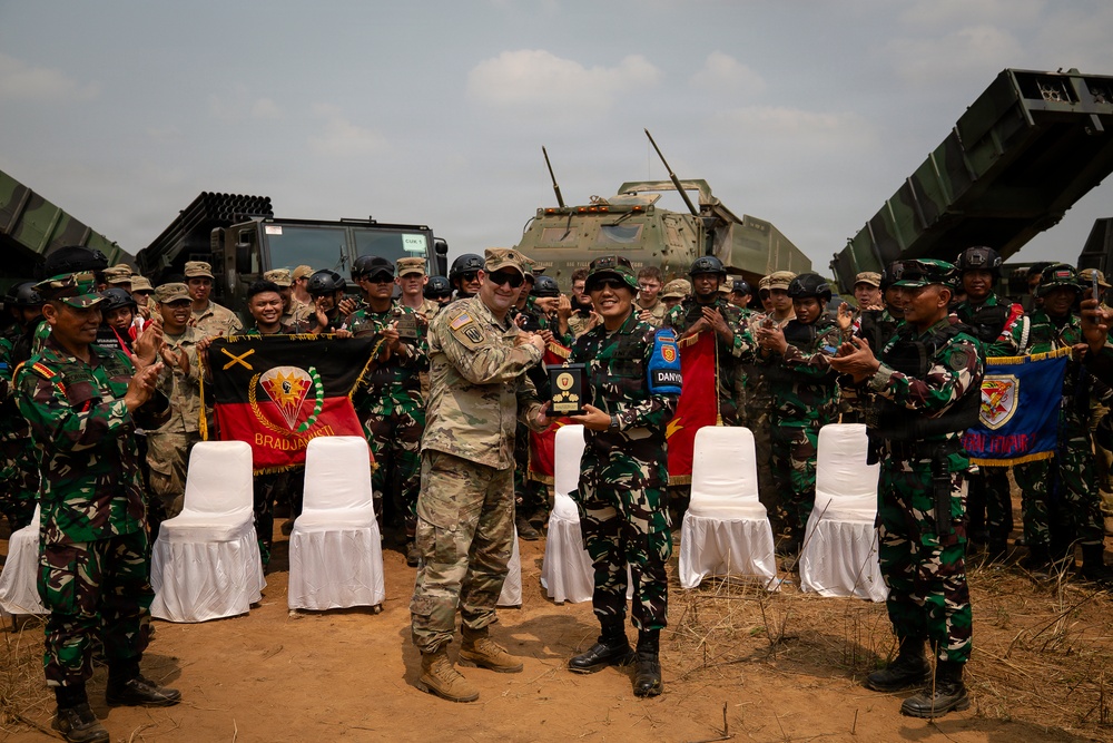 A military ceremony appears to be taking place between U.S. Army personnel and Indonesian Armed Forces (TNI) soldiers, with two central figures exchanging a plaque or award. The group is assembled in front of military vehicles, including what appears to be a HIMARS or similar rocket artillery system. A unit banner reading 'BRADJAJISTI' is visible among the gathered troops.