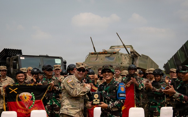 A military ceremony appears to be taking place between U.S. Army personnel and Indonesian Armed Forces (TNI) soldiers, with two central figures exchanging a plaque or award. The group is assembled in front of military vehicles, including what appears to be a HIMARS or similar rocket artillery system. A unit banner reading 'BRADJAJISTI' is visible among the gathered troops.