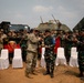 A military ceremony appears to be taking place between U.S. Army personnel and Indonesian Armed Forces (TNI) soldiers, with two central figures exchanging a plaque or award. The group is assembled in front of military vehicles, including what appears to be a HIMARS or similar rocket artillery system. A unit banner reading 'BRADJAJISTI' is visible among the gathered troops.