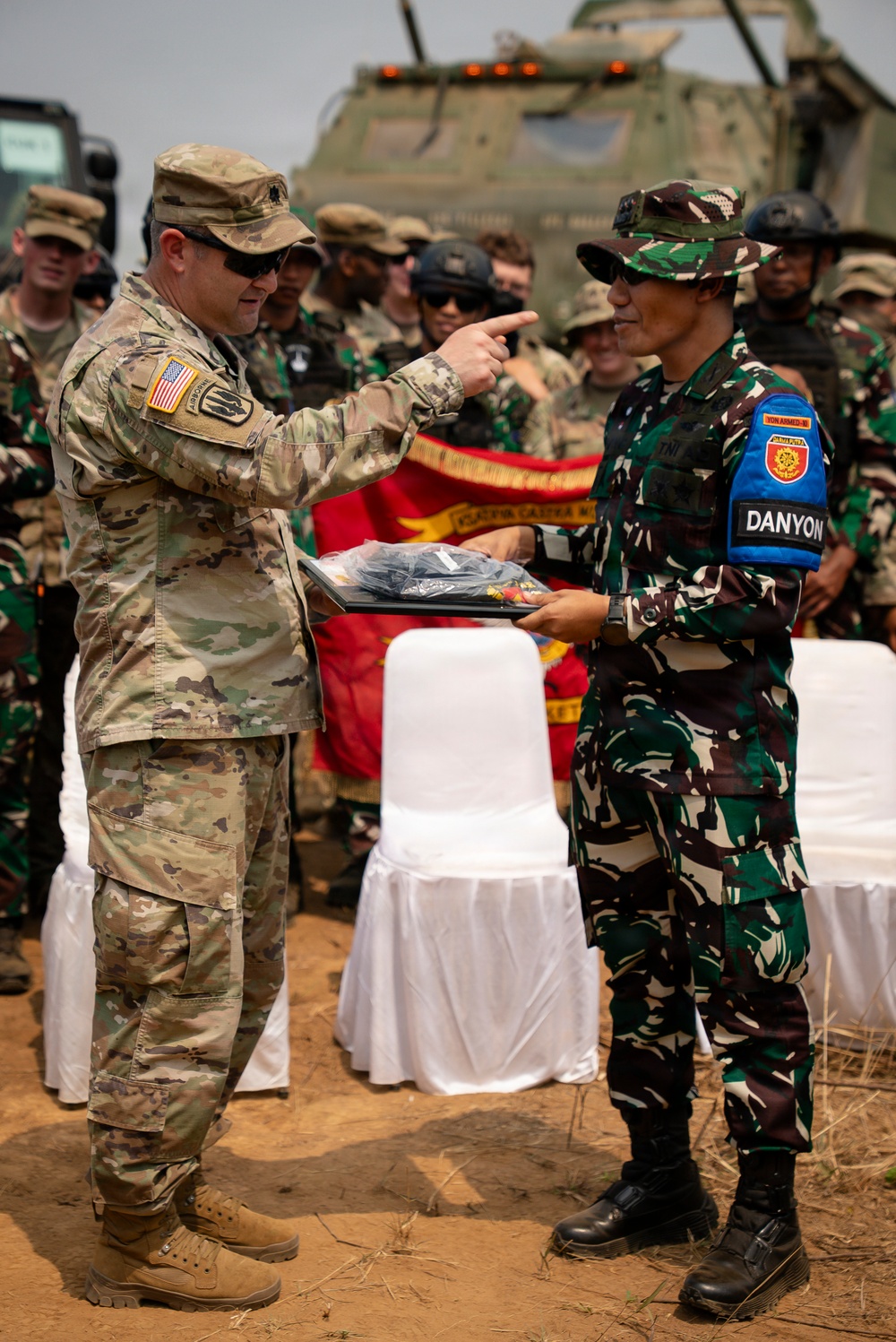 Two military personnel from different nations are engaged in what appears to be a formal exchange or presentation ceremony outdoors. A U.S. Army soldier wearing Airborne insignia and an American flag patch is pointing toward an Indonesian Army (TNI) soldier wearing a 'DANYON' armband, while both hold a presentation tray with items. Military personnel from both nations are gathered in the background near armored vehicles.