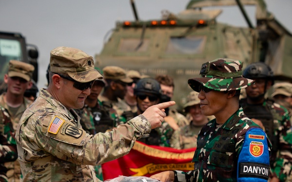 Two military personnel from different nations are engaged in what appears to be a formal exchange or presentation ceremony outdoors. A U.S. Army soldier wearing Airborne insignia and an American flag patch is pointing toward an Indonesian Army (TNI) soldier wearing a 'DANYON' armband, while both hold a presentation tray with items. Military personnel from both nations are gathered in the background near armored vehicles.