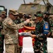 Two military personnel from different nations are engaged in what appears to be a formal exchange or presentation ceremony outdoors. A U.S. Army soldier wearing Airborne insignia and an American flag patch is pointing toward an Indonesian Army (TNI) soldier wearing a 'DANYON' armband, while both hold a presentation tray with items. Military personnel from both nations are gathered in the background near armored vehicles.