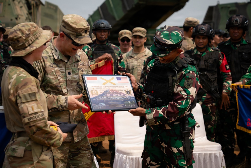 A military ceremony showing U.S. Army personnel presenting a commemorative plaque to an Indonesian military officer. The plaque features both the Indonesian and American flags, suggesting a joint military exercise or partnership event. Multiple soldiers from both nations are visible in the background.