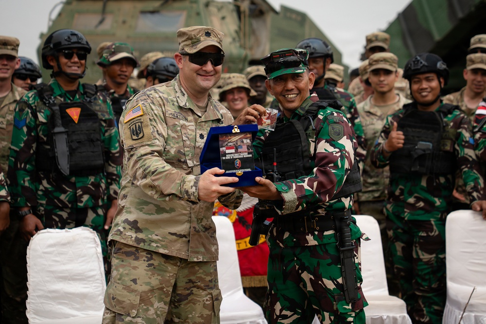 Two military officers — one in US Army multicam uniform and one in Indonesian Armed Forces (TNI) green camouflage — are exchanging a ceremonial plaque or commemorative gift during what appears to be a joint military exercise or bilateral event. A large group of soldiers from both nations stands in the background, many smiling, with at least one giving a thumbs-up. A military armored vehicle is visible in the background.