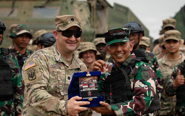 Two military officers — one in US Army multicam uniform and one in Indonesian Armed Forces (TNI) green camouflage — are exchanging a ceremonial plaque or commemorative gift during what appears to be a joint military exercise or bilateral event. A large group of soldiers from both nations stands in the background, many smiling, with at least one giving a thumbs-up. A military armored vehicle is visible in the background.