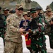 Two military officers — one in US Army multicam uniform and one in Indonesian Armed Forces (TNI) green camouflage — are exchanging a ceremonial plaque or commemorative gift during what appears to be a joint military exercise or bilateral event. A large group of soldiers from both nations stands in the background, many smiling, with at least one giving a thumbs-up. A military armored vehicle is visible in the background.