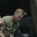 A medical provider wearing a stethoscope and blue nitrile gloves is attending to a patient lying supine on what appears to be a litter or stretcher. The provider is wearing an olive/tan military-style t-shirt and has visible tattoos on both arms. The setting appears to be a field medical or tactical environment based on the equipment visible in the background.
