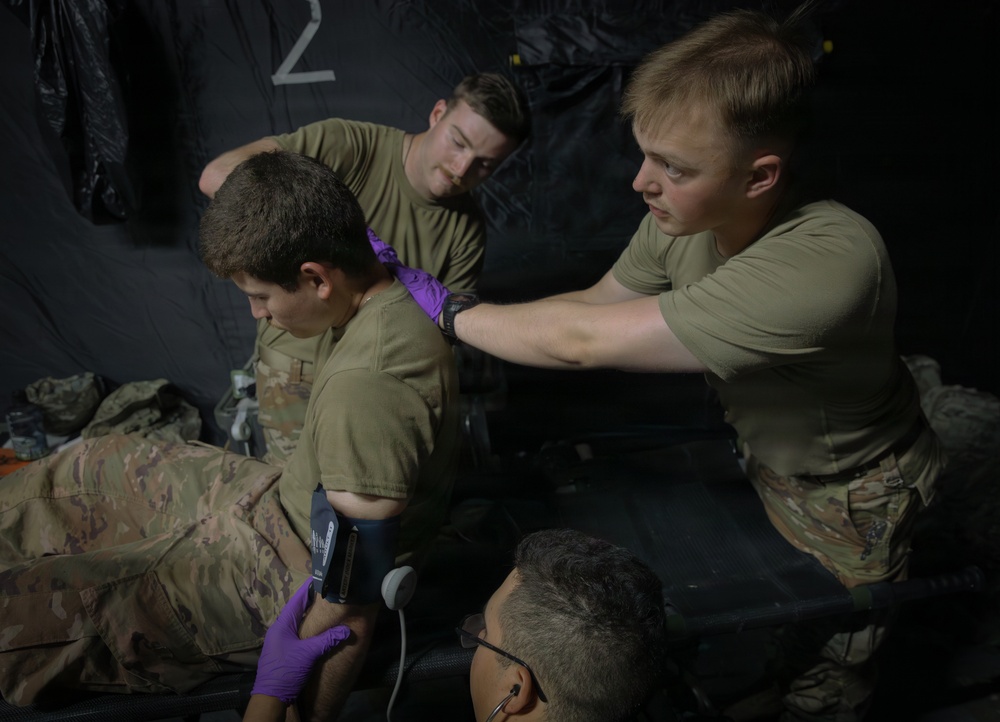 Military personnel in olive drab uniforms are conducting what appears to be a medical training exercise inside a dark tent or field medical station. Two soldiers are attending to a seated patient/casualty, with one applying what appears to be a cervical collar or neck dressing while another holds an ultrasound probe against the patient's torso. A fourth individual wearing glasses is visible in the foreground observing the procedure.