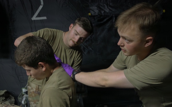 Military personnel in olive drab uniforms are conducting what appears to be a medical training exercise inside a dark tent or field medical station. Two soldiers are attending to a seated patient/casualty, with one applying what appears to be a cervical collar or neck dressing while another holds an ultrasound probe against the patient's torso. A fourth individual wearing glasses is visible in the foreground observing the procedure.