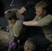 Military personnel in olive drab uniforms are conducting what appears to be a medical training exercise inside a dark tent or field medical station. Two soldiers are attending to a seated patient/casualty, with one applying what appears to be a cervical collar or neck dressing while another holds an ultrasound probe against the patient's torso. A fourth individual wearing glasses is visible in the foreground observing the procedure.