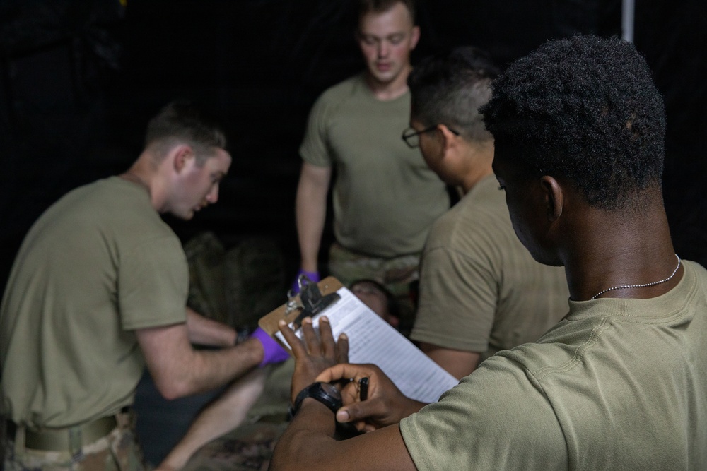 The image shows a group of military personnel in olive drab t-shirts engaged in what appears to be a medical training exercise. One individual is wearing purple medical gloves and appears to be performing a procedure or assessment on a person lying down, while another holds a clipboard with a checklist. The scene is set in a low-light environment consistent with a field training scenario.